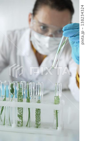 Woman scientist with blue gloves, face mask and protective glasses, is holding a test tube with plants inside in laboratory, vertical portrait view. Science and medicine concept 138234324