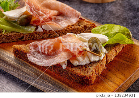 Rustic rye bread toasts with cured ham, cheese, and lettuce on a wooden cutting board. 138234931