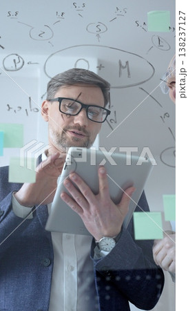 Focused businessman analyzing financial data and brainstorming new project strategies, writing important notes on a transparent glass board wall with sticky notes while holding a tablet Focused businessman analyzing financial data and brainstorming new project strategies, writing important notes on a transparent glass board wall with sticky notes while holding a tablet 138237127
