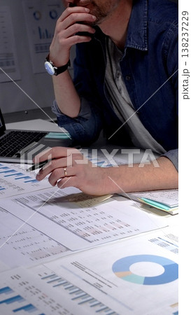 Caucasian man sitting at an office desk in the evening, concentrating and writing notes while surrounded by business documents, charts, graphs, and a laptop Caucasian man sitting at an office desk in the evening, concentrating and writing notes while surrounded by business documents, charts, graphs, and a laptop 138237229