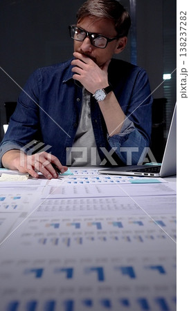 Professional man in glasses thinking deeply while analyzing financial charts and data on an office desk, working late night in a dark setting, feeling focused and determined 138237282