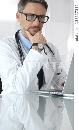 Male doctor wearing spectacles, thinking about healthcare diagnosis while using a laptop in a clinic office. Medicine and health care Male doctor wearing spectacles, thinking about healthcare diagnosis while using a laptop in a clinic office. Medicine and health care 138237356