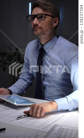 Businessman working late analyzing financial data at office, holding tablet with charts, writing notes on documents. Audit and taxes in business Businessman working late analyzing financial data at office, holding tablet with charts, writing notes on documents. Audit and taxes in business 138237384