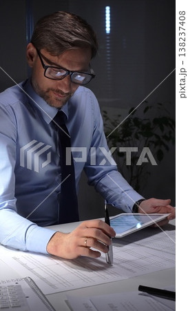 Businessman working late analyzing financial data at office, holding tablet with charts, writing notes on documents. Audit and taxes in business Businessman working late analyzing financial data at office, holding tablet with charts, writing notes on documents. Audit and taxes in business 138237408
