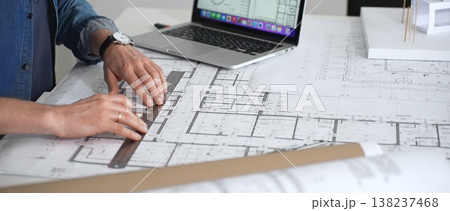 Architect sketching building plans on paper using a ruler and pen while working digitally on a laptop with a blueprint software displaying a floor plan Architect sketching building plans on paper using a ruler and pen while working digitally on a laptop with a blueprint software displaying a floor plan 138237468