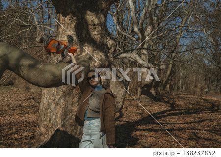 Lifestyle portrait of a stylish young woman in a cozy brown coat, light baggy jeans, holding a small dog in a orange sweater on a tree branch in a sunny spring park. Pet care and bond, outdoor leisure Lifestyle portrait of a stylish young woman in a cozy brown coat, light baggy jeans, holding a small dog in a orange sweater on a tree branch in a sunny spring park. Pet care and bond, outdoor leisure 138237852