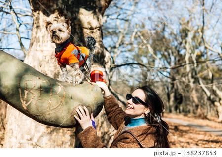 Candid lifestyle portrait of a stylish woman in a wool coat bonding with her small dog in a vibrant orange vest on a massive tree branch in a sun-drenched spring park. Emotional pet-owner connection. Candid lifestyle portrait of a stylish woman in a wool coat bonding with her small dog in a vibrant orange vest on a massive tree branch in a sun-drenched spring park. Emotional pet-owner connection. 138237853