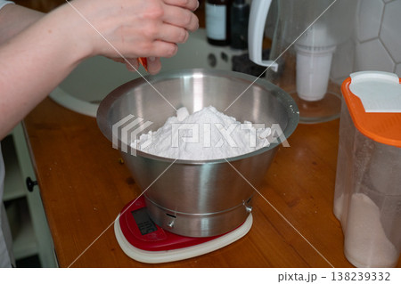 Hand adding ingredient into flour in metal bowl on kitchen scale, baking preparation on wooden countertop in home kitchen. Baking content, recipe illustration, cooking education, culinary blog, food 138239332