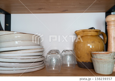 Ceramic jug, glass cups, pepper mill and wooden container on kitchen shelf, neutral kitchen storage arrangement with wooden surface. Kitchen organization, home interior, cooking content 138239357