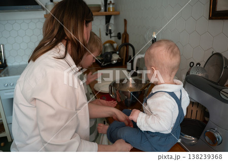Mother prepares ingredients while baby sits on kitchen countertop during home baking routine. Parenting, family cooking, child involvement, domestic lifestyle, cozy home 138239368