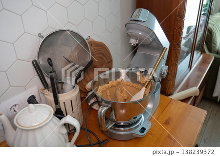 Stand mixer with dough in metal bowl on wooden countertop, kitchen utensils and kettle near tiled wall, home baking preparation scene. Cooking content, recipe illustration, kitchen workflow 138239372