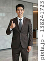Confident handsome young Asian businessman in suit and tie giving thumbs up gesture, smiling in a modern office hallway interior. 138241723