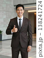 Confident handsome young Asian businessman in suit and tie giving thumbs up gesture, smiling in a modern office hallway interior. 138241724