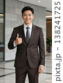 Confident handsome young Asian businessman in suit and tie giving thumbs up gesture, smiling in a modern office hallway interior. 138241725