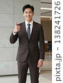 Confident handsome young Asian businessman in suit and tie giving thumbs up gesture, smiling in a modern office hallway interior. 138241726
