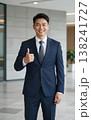 Confident handsome young Asian businessman in suit and tie giving thumbs up gesture, smiling in a modern office hallway interior. 138241727