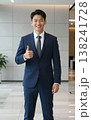 Confident handsome young Asian businessman in suit and tie giving thumbs up gesture, smiling in a modern office hallway interior. 138241728