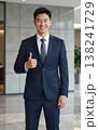Confident handsome young Asian businessman in suit and tie giving thumbs up gesture, smiling in a modern office hallway interior. 138241729