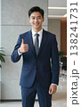 Confident handsome young Asian businessman in suit and tie giving thumbs up gesture, smiling in a modern office hallway interior. 138241731