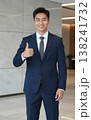 Confident handsome young Asian businessman in suit and tie giving thumbs up gesture, smiling in a modern office hallway interior. 138241732