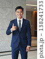 Confident handsome young Asian businessman in suit and tie giving thumbs up gesture, smiling in a modern office hallway interior. 138241733