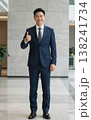 Confident handsome young Asian businessman in suit and tie giving thumbs up gesture, smiling in a modern office hallway interior. 138241734