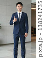Confident handsome young Asian businessman in suit and tie giving thumbs up gesture, smiling in a modern office hallway interior. 138241735