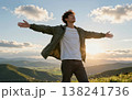 Handsome young Asian man spreading his arms wide open in joy at sunset, wearing a jacket and jeans amidst green hills and majestic mountains. 138241736