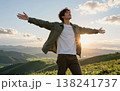Handsome young Asian man spreading his arms wide open in joy at sunset, wearing a jacket and jeans amidst green hills and majestic mountains. 138241737