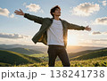 Handsome young Asian man spreading his arms wide open in joy at sunset, wearing a jacket and jeans amidst green hills and majestic mountains. 138241738