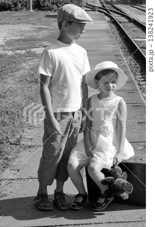Two children in vintage clothing wait on a train platform with a suitcase and teddy bear Two children in vintage clothing wait on a train platform with a suitcase and teddy bear 138241923