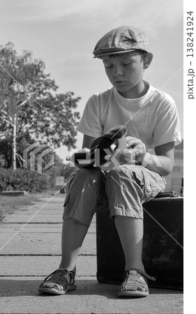 Young boy in flat cap sits on suitcase holding a stuffed toy, looking thoughtful Young boy in flat cap sits on suitcase holding a stuffed toy, looking thoughtful 138241924