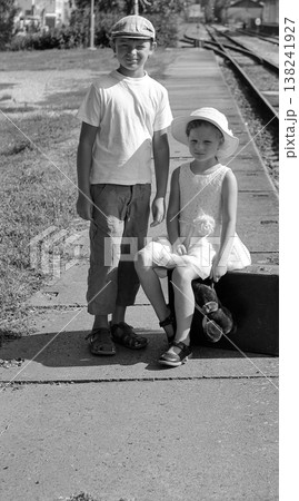 Two children, a boy and a girl, wait on a train platform with a suitcase and teddy bear Two children, a boy and a girl, wait on a train platform with a suitcase and teddy bear 138241927