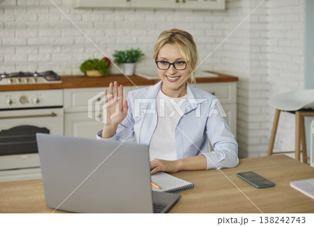 Happy young woman sitting at wooden desk in home kitchen at laptop online, looking at camera, waving Happy young woman sitting at wooden desk in home kitchen at laptop online, looking at camera, waving 138242743
