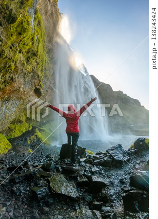 Visitor stands with arms raised in excitement behind Seljalandsfoss Waterfall in Iceland. The sun sets, illuminating the lush landscape and cascading water. Visitor stands with arms raised in excitement behind Seljalandsfoss Waterfall in Iceland. The sun sets, illuminating the lush landscape and cascading water. 138243424