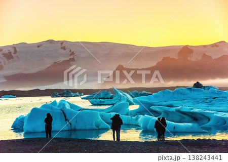 Tourists admire the stunning sunset over Iceland's Glacier Lagoon, surrounded by large icebergs. The serene scene captures the beauty of nature 138243441