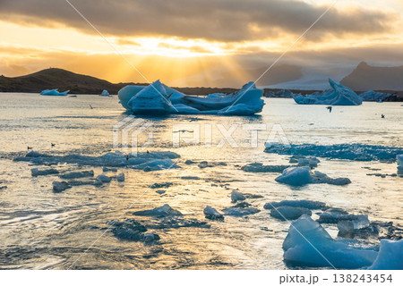 Icebergs float in Iceland's glacier lagoon, reflecting the warm colors of sunset. Birds fly above, adding life to the tranquil scene. This location is a must-see for nature enthusiasts. Icebergs float in Iceland's glacier lagoon, reflecting the warm colors of sunset. Birds fly above, adding life to the tranquil scene. This location is a must-see for nature enthusiasts. 138243454