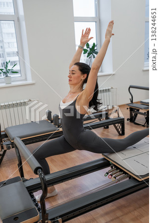 Athletic woman raising both arms up practicing pilates on reformer machine near windows. Reformer machine, arms up, pilates, windows, athletic. Athletic woman raising both arms up practicing pilates on reformer machine near windows. Reformer machine, arms up, pilates, windows, athletic. 138244651