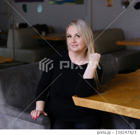 Elegant mature woman in black dress sitting on chair restaurant posing confident glamour lifestyle indoor sophisticated fashion 138247823