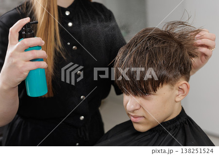 Hairdresser applies styling spray to a boy hair after a haircut. Close-up of hairstyling process, grooming routine, and finishing service in salon. Hairdresser applies styling spray to a boy hair after a haircut. Close-up of hairstyling process, grooming routine, and finishing service in salon. 138251524