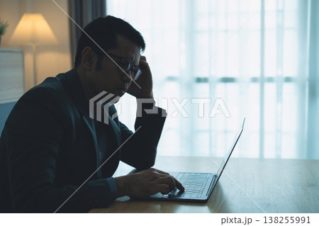 Businessman in Dark Suit Worrying Over Laptop While Working Late Night in Modern Home Office with Soft Light from Window Businessman in Dark Suit Worrying Over Laptop While Working Late Night in Modern Home Office with Soft Light from Window 138255991
