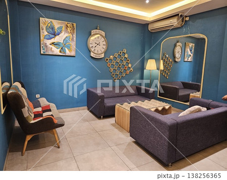 A stylish modern living room interior featuring purple fabric sofas, a unique wavy wooden coffee table, and a large vintage wall clock against deep teal walls with a tall arched gold-framed mirror. A stylish modern living room interior featuring purple fabric sofas, a unique wavy wooden coffee table, and a large vintage wall clock against deep teal walls with a tall arched gold-framed mirror. 138256365