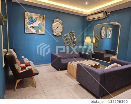 A stylish modern living room interior featuring purple fabric sofas, a unique wavy wooden coffee table, and a large vintage wall clock against deep teal walls with a tall arched gold-framed mirror. A stylish modern living room interior featuring purple fabric sofas, a unique wavy wooden coffee table, and a large vintage wall clock against deep teal walls with a tall arched gold-framed mirror. 138256366