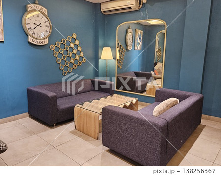 A stylish modern living room interior featuring purple fabric sofas, a unique wavy wooden coffee table, and a large vintage wall clock against deep teal walls with a tall arched gold-framed mirror. A stylish modern living room interior featuring purple fabric sofas, a unique wavy wooden coffee table, and a large vintage wall clock against deep teal walls with a tall arched gold-framed mirror. 138256367
