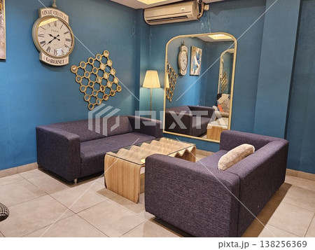 A stylish modern living room interior featuring purple fabric sofas, a unique wavy wooden coffee table, and a large vintage wall clock against deep teal walls with a tall arched gold-framed mirror. 138256369