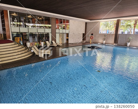 A modern luxury indoor swimming pool featuring crystal clear blue water, elegant wooden decking with sun loungers, and a stylish glass-walled fitness center in a high-end hotel or resort area. 138260050