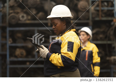 Hispanic female foreman and black female worker standing holding tablet, both of them checking inventory of auto parts and engine maintenance duties in factory. Hispanic female foreman and black female worker standing holding tablet, both of them checking inventory of auto parts and engine maintenance duties in factory. 138261551
