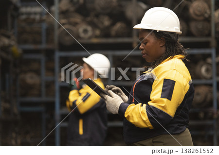 Hispanic female foreman and black female worker standing holding tablet, both of them checking inventory of auto parts and engine maintenance duties in factory. Hispanic female foreman and black female worker standing holding tablet, both of them checking inventory of auto parts and engine maintenance duties in factory. 138261552