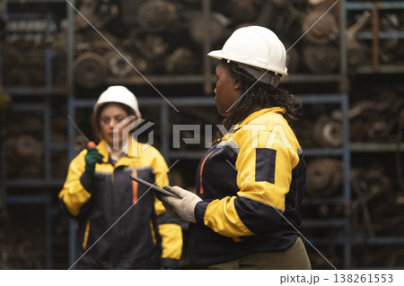 Hispanic female foreman and black female worker standing holding tablet, both of them checking inventory of auto parts and engine maintenance duties in factory. Hispanic female foreman and black female worker standing holding tablet, both of them checking inventory of auto parts and engine maintenance duties in factory. 138261553