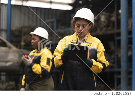 Hispanic female foreman and black female worker standing holding tablet, both of them checking inventory of auto parts and engine maintenance duties in factory. Hispanic female foreman and black female worker standing holding tablet, both of them checking inventory of auto parts and engine maintenance duties in factory. 138261557
